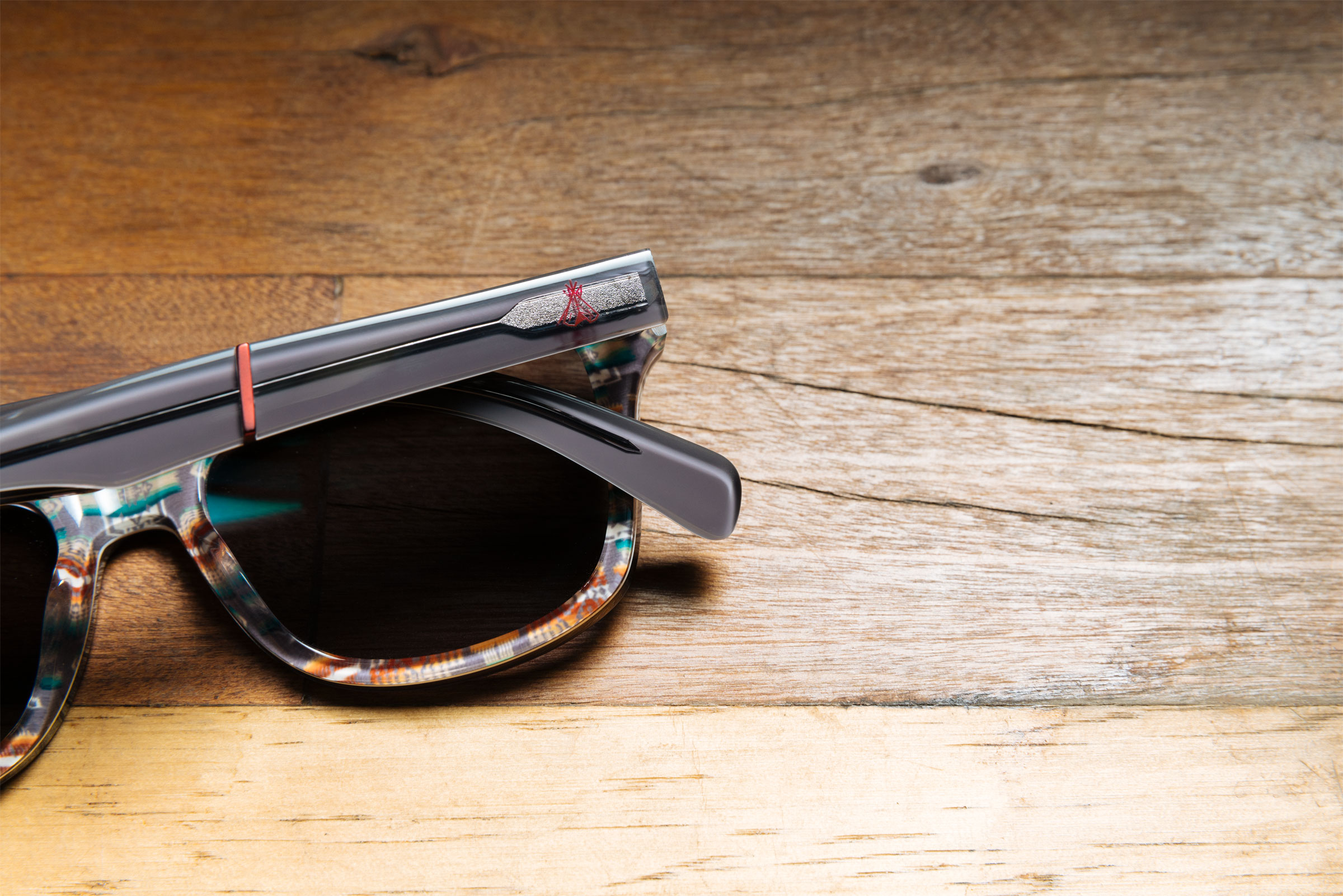 A pair of Pendleton x Shwood sunglasses sits on a wood table. You can see the details of the branding in this shot, including the Chief Joseph pattern and a small teepee in red on the eyeglass stem.