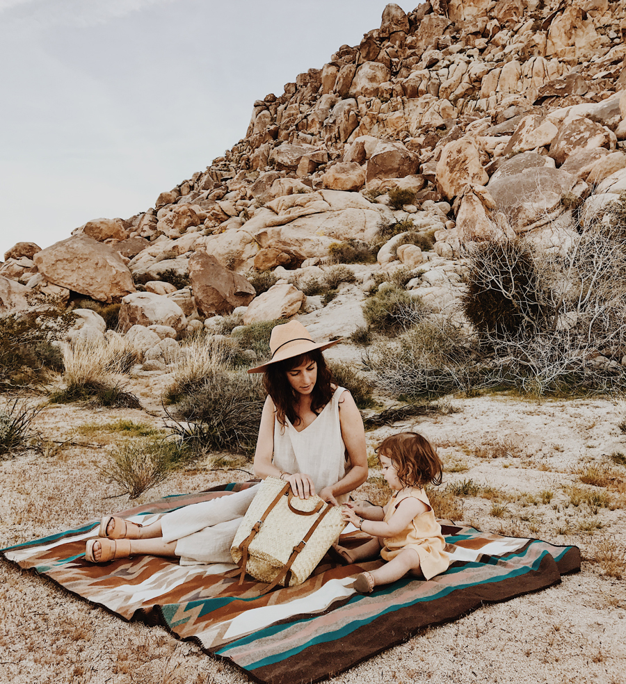 A mother and her young toddler daughter sit on a Pendleton Weavers Series blanket , on dry grass near a red rock hill.