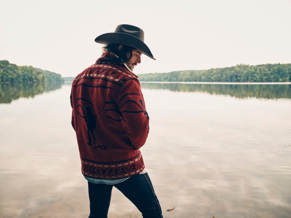 A man stands at a lakeshore wearing a hat and a Pendleton-Wrangler jacket. 