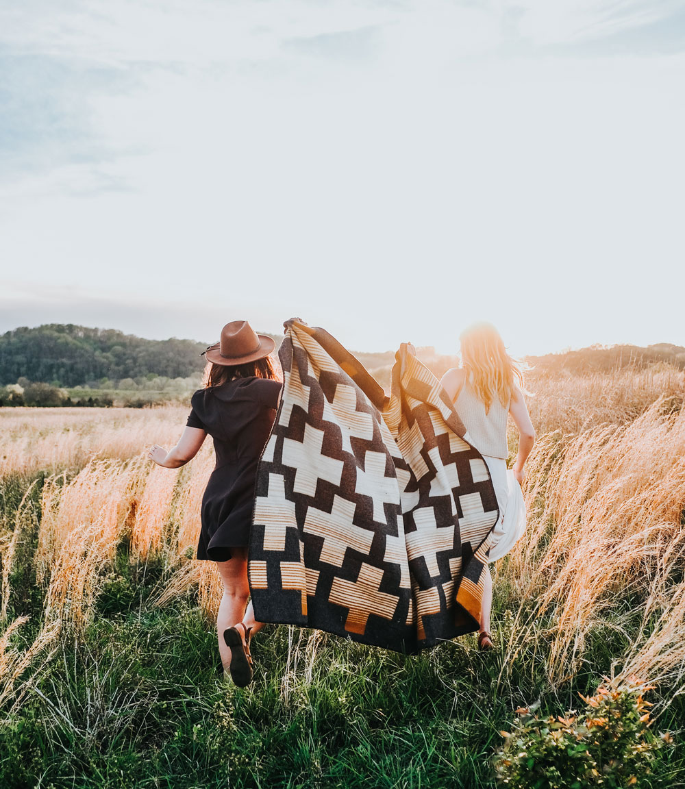 Two young women run away from the camera through a field with tall grasses in the sunshine. They are holding a pendleton Compass point blanket between them as they run.