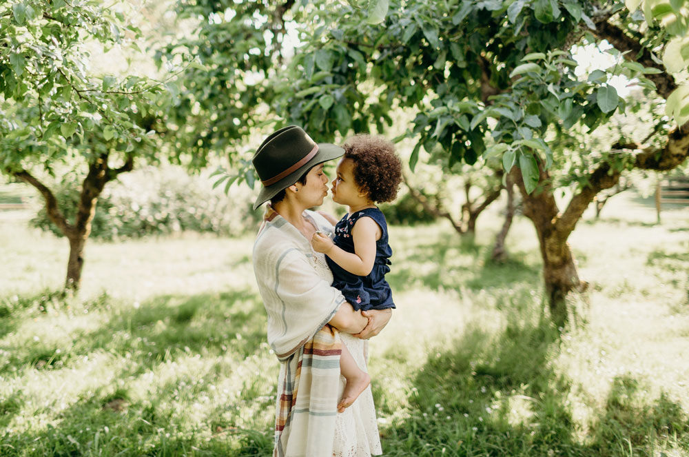A mother wearing a Pendleton scarf and hat, with her 2 year old daughter in a sun dappled orchard