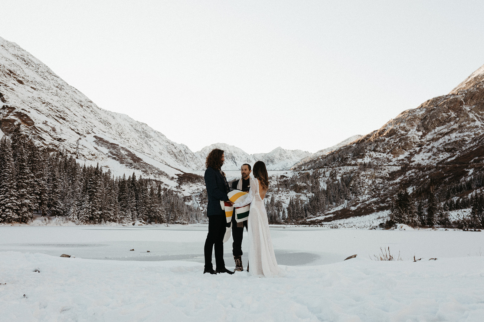 A man and woman hold a Pendleton blanket in the snow, while their officiant marries them. 