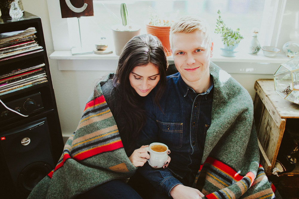 blonde man and brunette woman seated in front of a window, wrapped in a pendleton Camp Stripe blanket. Woman is holding a cup of coffee.