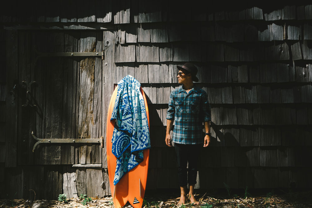 A young man stands next to an orange surfboard in front of a shingled wall. He is wearing a Pendleton Board Shirt in the Original Surf Plaid made famous by The Beach Boys.