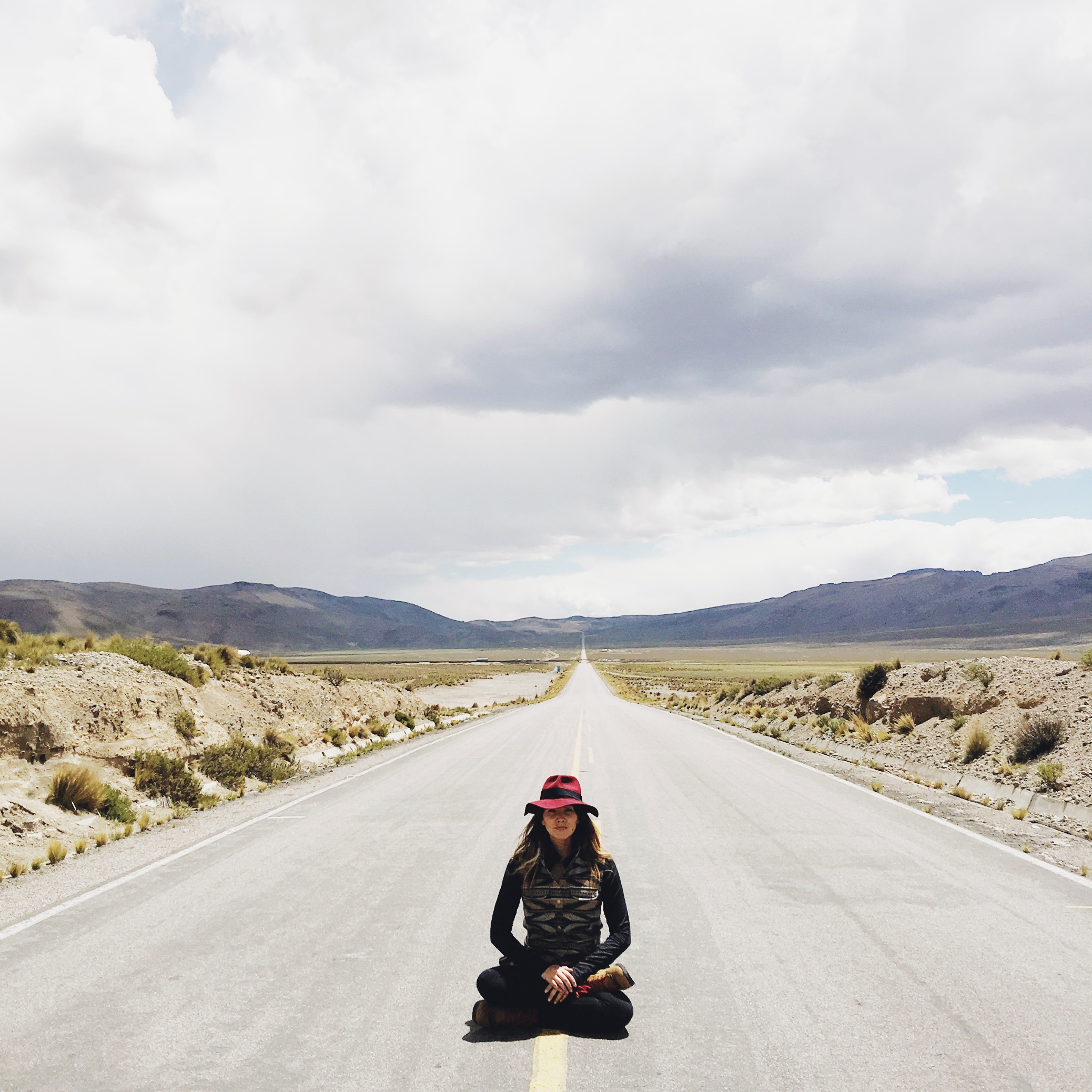 A woman sits crosslegged on a highway, with the road receding into the far distance.