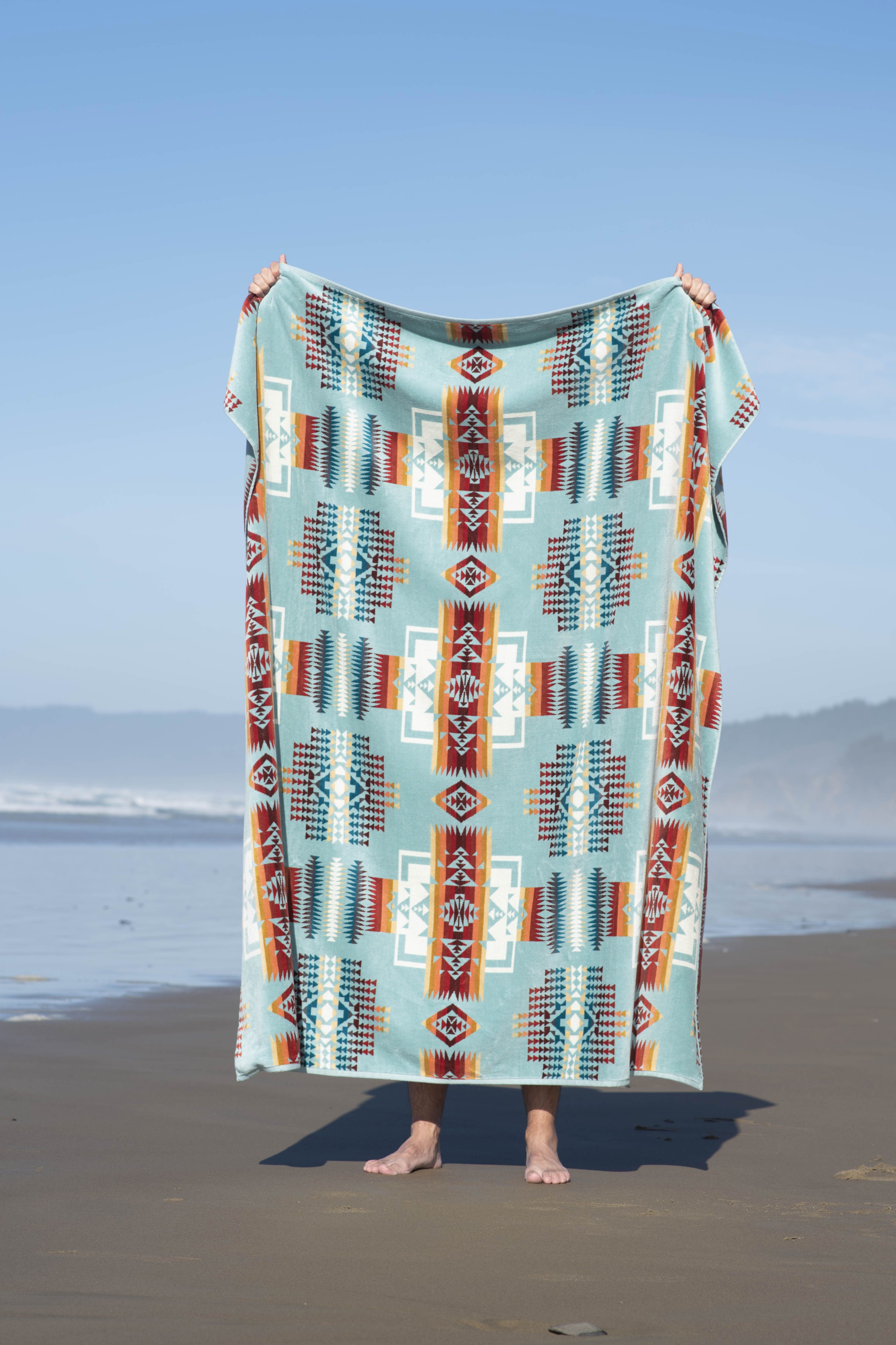 A person stands on a sandy beach holding up a Pendleton Chief Joseph towel.